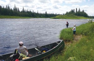 Guide Joshua Sutherland and his canoe rest on a scenic Ontario river bank while two of his clients fish from the bank. 